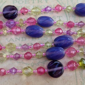 May include: Close-up of several strands of colourful beaded necklaces. The beads are in shades of purple, pink, green, and yellow. The beads are various shapes, including round, oval, and faceted. The necklaces are strung on a pink string.