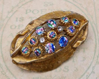 May include: A gold-toned metal brooch shaped like an elongated leaf with a textured surface. The center of the brooch is open and features a cluster of round, iridescent, multi-colored rhinestones that display blue, purple, and pink hues.