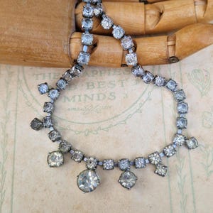 May include: A vintage silver necklace adorned with clear, sparkling rhinestones. The necklace has a delicate chain with square and round-cut stones, and larger, dangling stones. The background includes the text "THE HAPPIEST BEST MINDS."