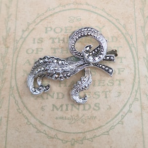 May include: A silver-toned brooch with a swirling, ornate design. The brooch features a bow shape with a central loop and flowing, curved elements. Small, sparkling embellishments are set throughout the design, adding a touch of glamour. The brooch is set against a vintage-style background.