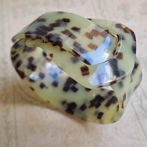 May include: A wide, curved bangle bracelet featuring a translucent pale green base with a scattered pattern of dark brown, rectangular-shaped spots. The bracelet has a smooth, glossy finish and a continuous, fluid shape that wraps around itself.