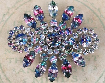 May include: A brooch featuring a central round iridescent crystal surrounded by smaller round and marquise-shaped crystals in shades of blue, pink, and purple. The stones are set in a silver-toned metal frame, creating a starburst pattern.
