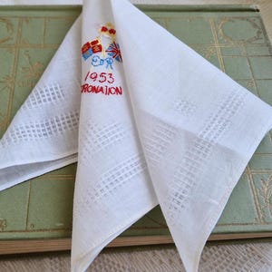 May include: White handkerchief with embroidered details, including a crown, flags, and the text "1953 CORONATION" in red. The handkerchief is folded and lies on a vintage green book.