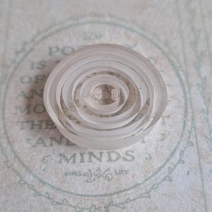 May include: A clear, circular button with a spiral design. The button has a series of concentric, raised rings that create a textured surface. Two small holes are visible in the center. This button is suitable for sewing projects.