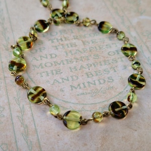 May include: A vintage necklace with green and brown glass beads of different sizes, connected by a gold-toned chain. The beads have a striped design. The background features faded text.