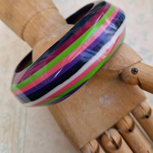 Könnte beinhalten: Ein farbenfrohes Armreif-Armband mit einem gestreiften Muster in Rosa, Grün, Lila, Schwarz und Weiß. Das glänzende Armband wird auf einer Holzhand präsentiert. Dieses Accessoire ist perfekt, um jedem Outfit einen Farbtupfer zu verleihen. Der Durchmesser beträgt ca. 6,5 cm.