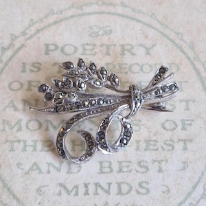 Op de afbeelding: Zilverkleurige broche in de vorm van een tarweoogst, versierd met kleine, donkere edelstenen. De broche heeft een strik en een speldsluiting. De achtergrond heeft vervaagde tekst die luidt "POETRY IS..."