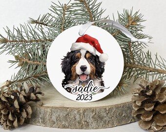 May include: White round Christmas bauble featuring a Bernese Mountain Dog wearing a red Santa hat. The dog's name "Sadie" and the year "2023" are written beneath the dog.
