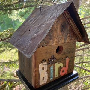 May include: A wooden birdhouse with a corrugated metal roof and the word "Bird" in colourful letters. The birdhouse has a round entry hole and a black base, suspended by a wire loop.