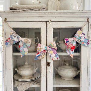 May include: A decorative garland featuring three fabric bows made from a patchwork of colorful floral and patterned cotton scraps. The bows are strung together on a thin, natural-colored jute twine, creating a festive hanging display.