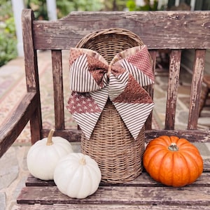 May include: A tall, cylindrical woven wicker basket adorned with a large, multi-patterned fabric bow. The bow features a mix of brown and tan striped, floral, and dotted prints. Two white pumpkins and one orange pumpkin sit at the base of the basket.