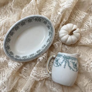 May include: A white ceramic oval dish with a green floral border, a white ceramic creamer with a green floral pattern and gold trim on the handle, and a small white ceramic pumpkin figurine with a gold stem, all arranged together.