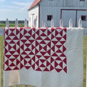 May include: A rectangular quilt featuring a repeating geometric pattern of deep red and off-white triangles. The central section displays a grid of square blocks composed of smaller triangles, while the bottom and right edges feature a solid border.