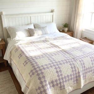 May include: A bed quilt featuring a repeating lavender and cream geometric chain pattern. The quilt is draped over a bed with white pillows and a white bedspread, showcasing a detailed stitched texture across the fabric surface.