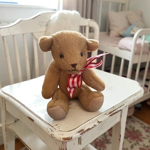 May include: A brown teddy bear with a red and white checkered ribbon around its neck. The bear is sitting on a white, distressed wooden stool. The bear has tan paws and a stitched nose and mouth.