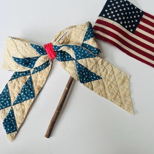 May include: A patriotic-themed decorative bow and American flag. The bow is made of quilted fabric with blue star patterns and a red center. The flag is red, white, and blue.