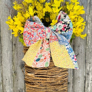 May include: A handmade spring wreath featuring a woven basket base, vibrant yellow flowers, and a large, colorful patchwork bow. The bow incorporates floral patterns in red, blue, and yellow fabrics, adding a touch of rustic charm.