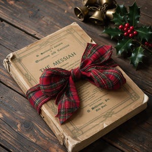May include: An antique book titled The Messiah with a worn tan cover, wrapped in a red and green plaid fabric bow. A sprig of green holly leaves with red berries and a cluster of small brass bells are placed near the top of the book.