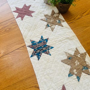 May include: Cream-colored quilted table runner with star-shaped patchwork designs in shades of red, brown, and blue. The runner is placed on a wooden table, with a teapot and potted plant visible in the background.