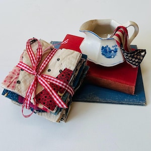 May include: A stack of fabric squares tied with a red and white checkered ribbon. A small white pitcher with a blue design and a red and white striped fabric is on top of two books, one red and one blue.