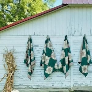 May include: Four square quilts featuring a repeating pattern of dark green solid blocks and beige blocks with a brown star design. The quilts are hung vertically against a white wooden wall, with two white pumpkins and dried corn stalks nearby.