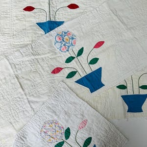 May include: A white quilted fabric piece featuring three appliquéd floral motifs. Each motif consists of a blue vase, green stems and leaves, and a large circular flower head made from patterned fabric, accompanied by smaller red or pink buds.