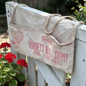 May include: A cream-colored canvas apron with two straps, featuring red printed text that reads True Value, Mahoney's Bldg. Supply, and TEL. 758-6921, along with a graphic of a person carrying lumber and the words Quality Building Supplies.