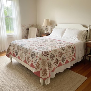 May include: A white bed frame holds a mattress covered in a quilt featuring a repeating geometric pattern in shades of red, brown, and tan. The quilt has a scalloped border and is topped with two white pillows. The bed is set in a room.