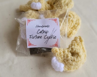 May include: Handmade catnip fortune cookies. The image shows several crocheted fortune cookies in a light yellow color with white accents. A clear plastic bag contains catnip and a label that reads "Handmade Catnip Fortune Cookie".
