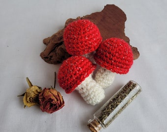 Peut inclure: Trois ornements en forme de champignon crochetés en rouge et blanc. Les champignons sont disposés sur un morceau de bois avec un petit flacon en verre d'herbes séchées.
