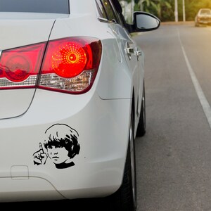May include: A white car with a black and white sticker of a person with short hair on the back bumper. The sticker is a silhouette of a person with their hand raised in a peace sign.