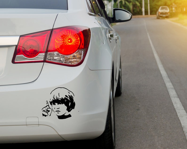 May include: A white car with a black and white sticker of a person with short hair on the back bumper. The sticker is a silhouette of a person with their hand raised in a peace sign.