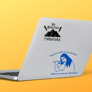 K&ouml;nnte beinhalten: Ein silberner Laptop mit zwei Aufklebern. Der obere Aufkleber ist schwarz und sagt "Big Mountain Fudgecake" mit einem schwarzen Berg und Blitzen. Der untere Aufkleber ist blau und sagt "There's a hole in my pocket where my money should go" mit einer Cartoonfigur, die ein Mikrofon h&auml;lt.