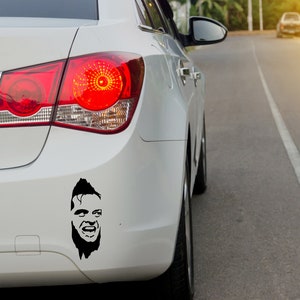 May include: A white car with a black and white sticker of a man's face on the back bumper.