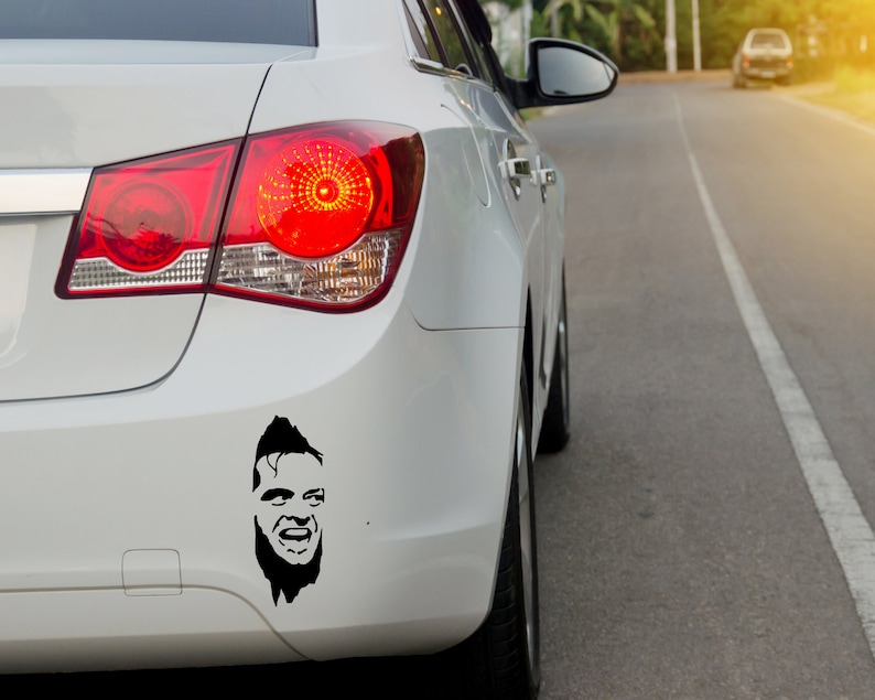 May include: A white car with a black and white sticker of a man's face on the back bumper.