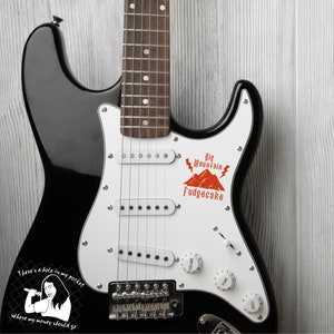 K&ouml;nnte beinhalten: Eine schwarze E-Gitarre mit einem wei&szlig;en Schlagbrett. Das Schlagbrett hat einen roten Aufkleber mit dem Text "Big Mountain Fudgecake" und einer Berggrafik. Die Gitarre hat auch einen Aufkleber auf dem unteren Korpus mit einer Comicfigur und dem Text "There's a hole in my pocket Where my money should go."