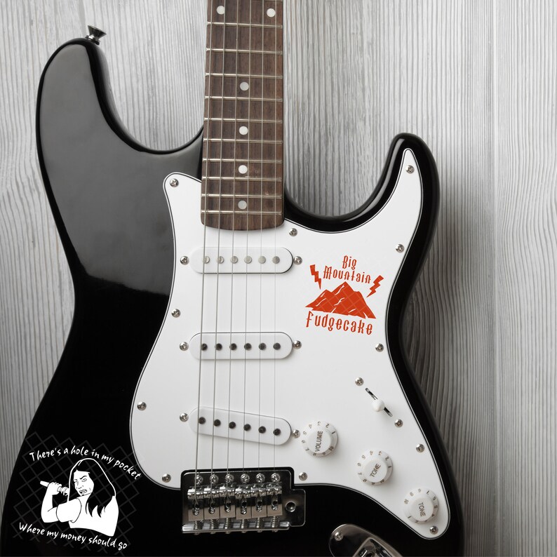 K&ouml;nnte beinhalten: Eine schwarze E-Gitarre mit einem wei&szlig;en Schlagbrett. Das Schlagbrett hat einen roten Aufkleber mit dem Text "Big Mountain Fudgecake" und einer Berggrafik. Die Gitarre hat auch einen Aufkleber auf dem unteren Korpus mit einer Comicfigur und dem Text "There's a hole in my pocket Where my money should go."