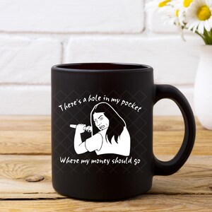 K&ouml;nnte beinhalten: Schwarze Keramik-Kaffeetasse mit einem wei&szlig;en Design. Das Design zeigt eine Frau mit langen Haaren, die ein Mikrofon h&auml;lt, und den Text "There's a hole in my pocket where my money should go."
