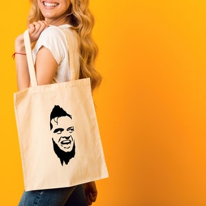 May include: A white tote bag with a black silhouette of a man's face on it. The man has a beard and is smiling.