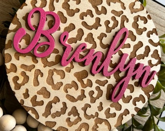 May include: A round wooden sign with a leopard print design. The name "Brielyn" is written in pink letters on the sign.