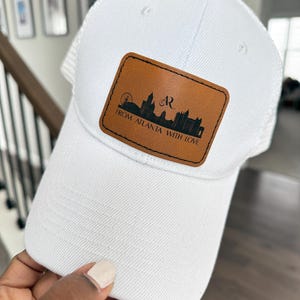 May include: White trucker hat with a brown leather patch featuring a black skyline graphic and the text "FROM ATLANTA WITH LOVE". The hat has a mesh back and a curved brim.