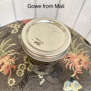 May include: A glass jar with a silver lid labelled "Ball Sure Tight" filled with a dark brown spice blend. The jar is sitting on a patterned surface with the text "Gowe from Mali" above it.