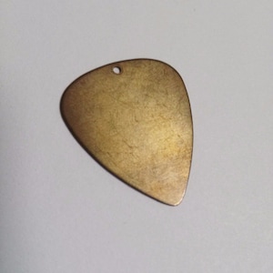 Puede incluir: Una púa de guitarra de color latón, con forma de triángulo redondeado y un pequeño agujero en la parte superior. La púa tiene una superficie texturizada y un aspecto ligeramente desgastado. Para músicos.