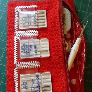 Könnte beinhalten: Eine rote Reißverschlusstasche mit weißen Punkten und einem weißen Rand. Die Tasche enthält vier Packungen Nähnadeln mit der Aufschrift "Schmetz" mit verschiedenen Nadeltypen und Größen. Die Tasche enthält außerdem einen weißen Fadenzieher und eine kleine weiße Spule.