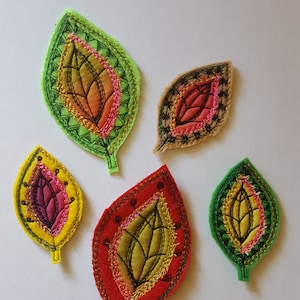 Może przedstawiać: Pięć haftowanych filcowych kształtów liści w różnych kolorach, w tym zielonym, żółtym, czerwonym i różowym. Każdy liść ma szczegółowy wzór z czarnymi ściegami i złotymi akcentami.