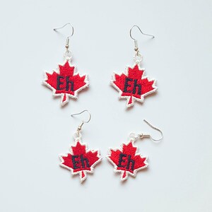 May include: Four pairs of earrings featuring a red maple leaf design with a white border. Each leaf has the word "Eh" embroidered in black. The earrings have silver-colored hooks.