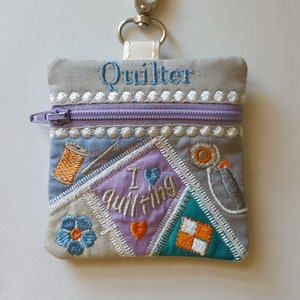 May include: A grey fabric zipped pouch with a patchwork design featuring a heart, a flower, and a sewing machine. The pouch has a silver key ring and the text "Quilter" and "I love quilting" embroidered on it.