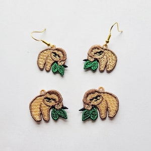 May include: A pair of gold dangle earrings featuring a brown sloth with green leaves. The sloth is embroidered and has a cute expression.