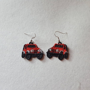 May include: A pair of embroidered earrings shaped like red and black jeeps. The earrings have silver-colored hooks and are set against a light background. The jeep details include headlights and tires.