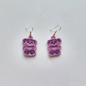 May include: A pair of lavender teddy bear earrings. Each earring features a detailed embroidered design of a teddy bear with silver-colored fish hook ear wires. The earrings are set against a white background.